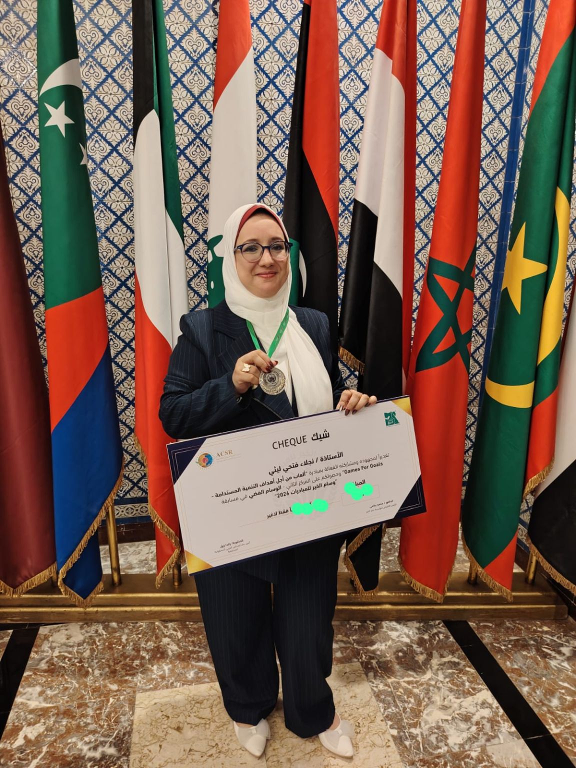 Une femme exhibant fièrement une médaille et un chèque, debout devant des drapeaux de la Ligue arabe